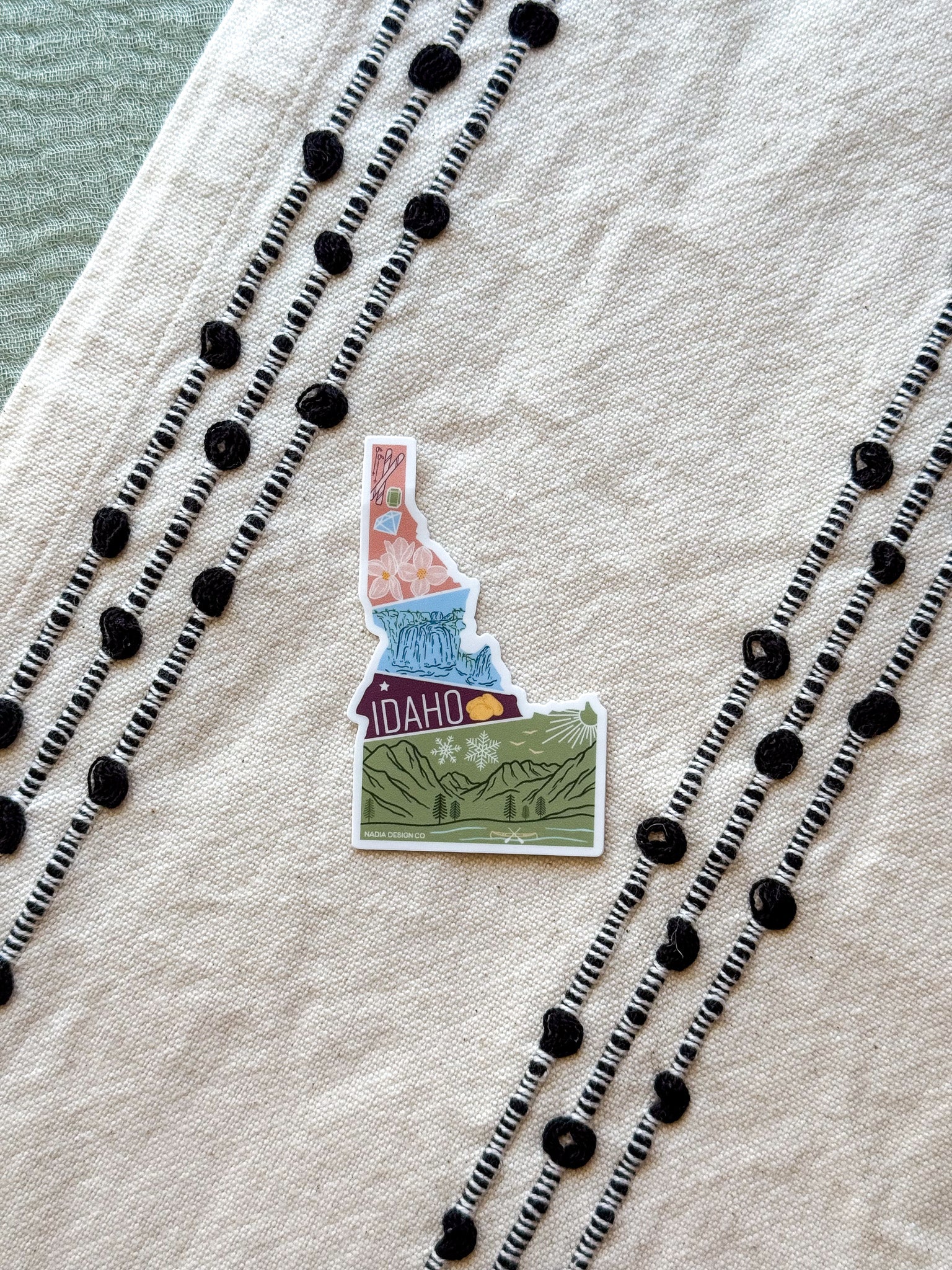 sticker of Idaho with scenic illustrations stamp design held by a hand on a textured fabric background