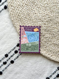 sticker of Idaho with scenic illustrations stamp design held by a hand on a textured fabric background