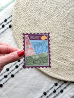 sticker of Idaho with scenic illustrations stamp design held by a hand on a textured fabric background