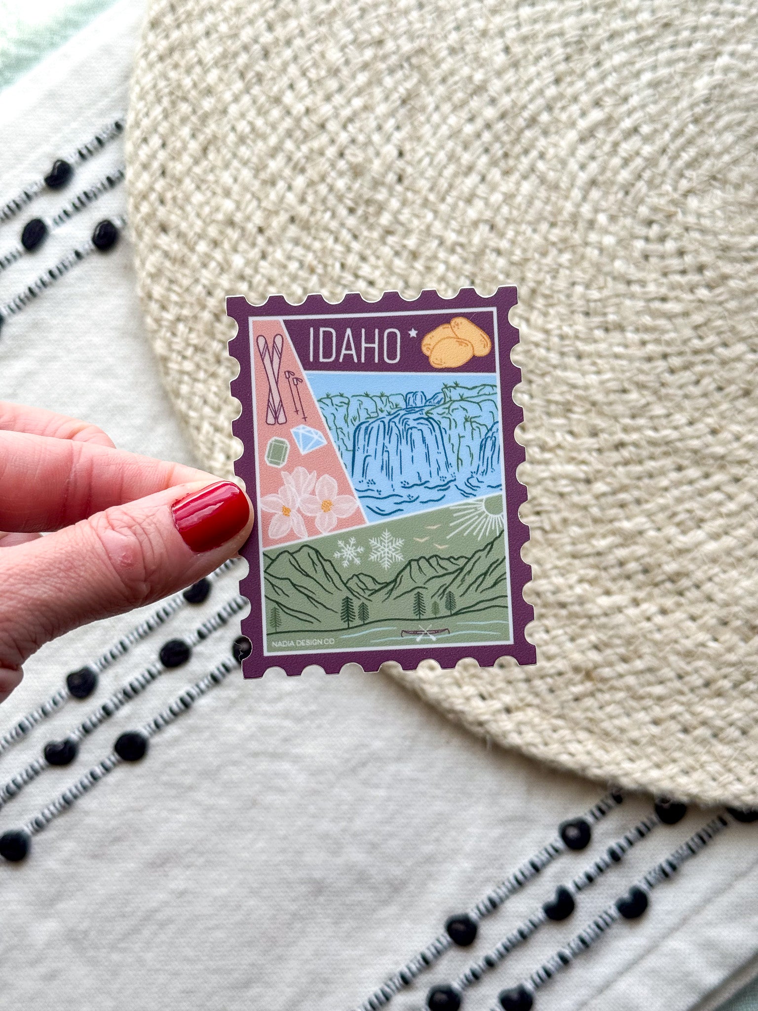 sticker of Idaho with scenic illustrations stamp design held by a hand on a textured fabric background