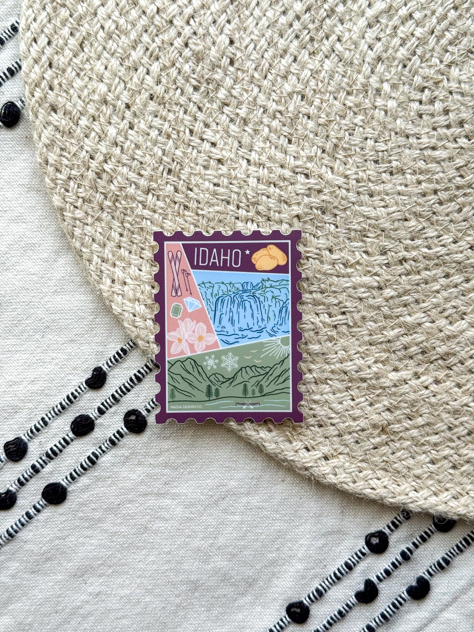 sticker of Idaho with scenic illustrations stamp design held by a hand on a textured fabric background