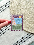 sticker of Idaho with scenic illustrations stamp design held by a hand on a textured fabric background