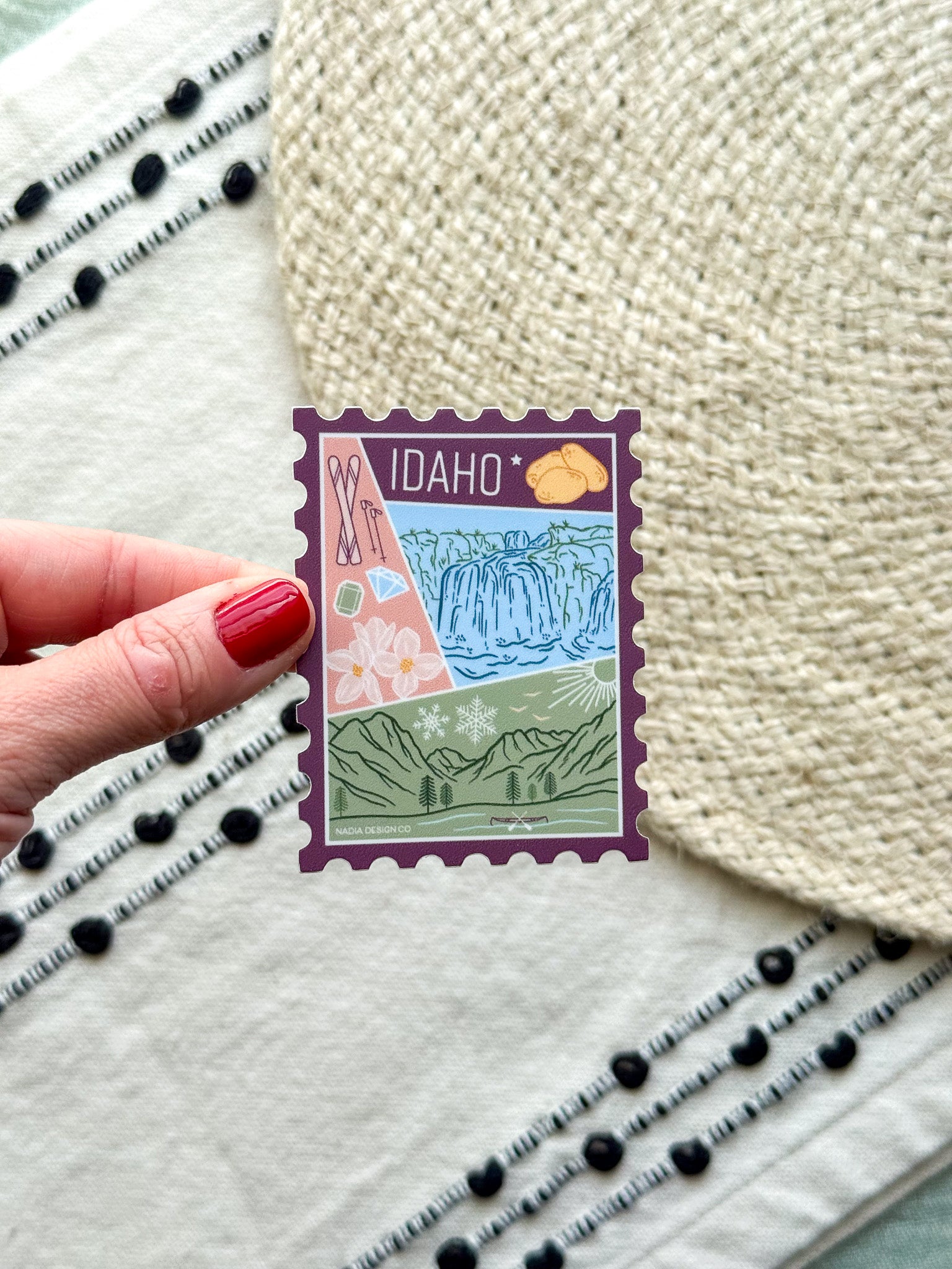 sticker of Idaho with scenic illustrations stamp design held by a hand on a textured fabric background