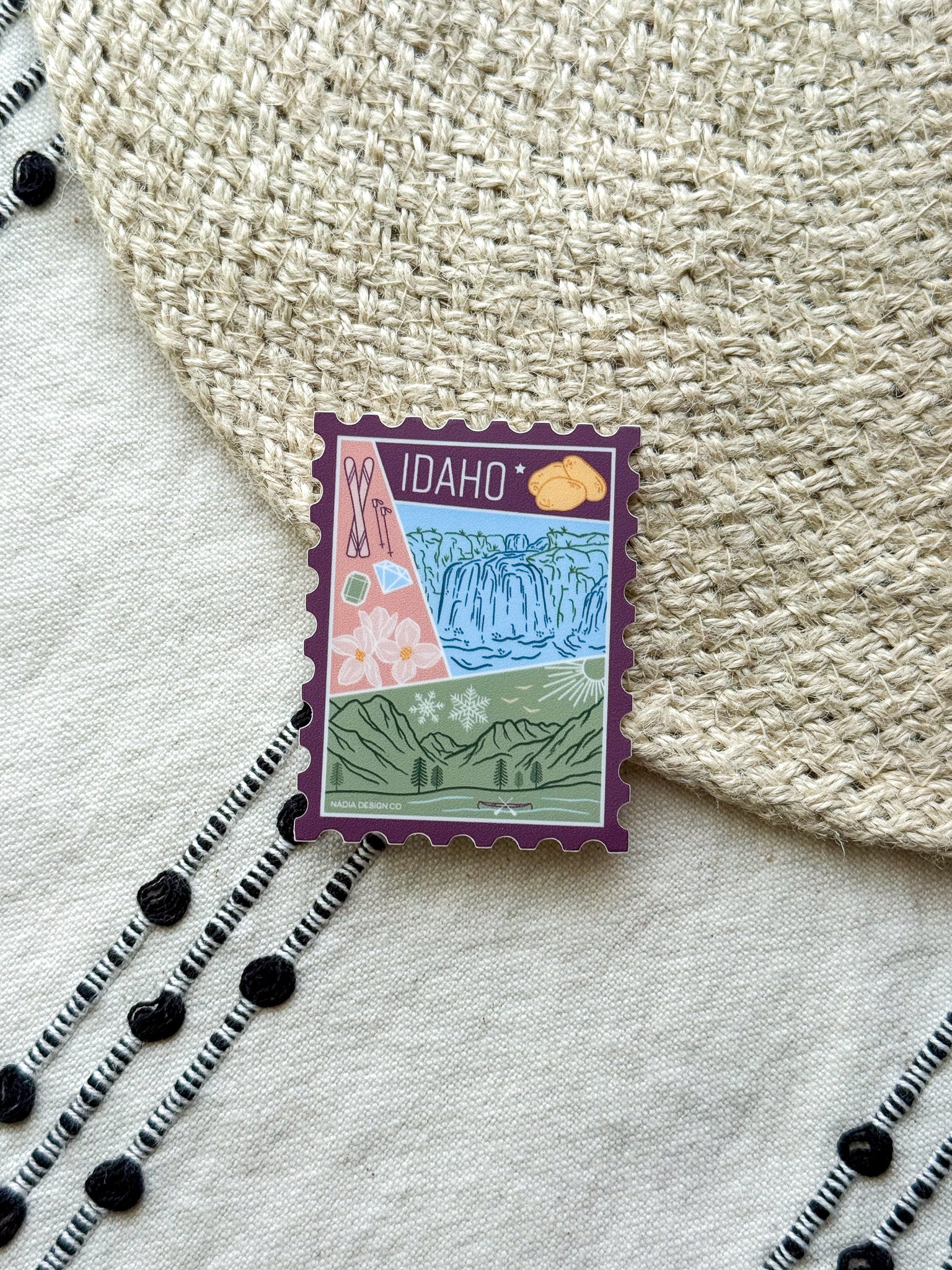 sticker of Idaho with scenic illustrations stamp design held by a hand on a textured fabric background