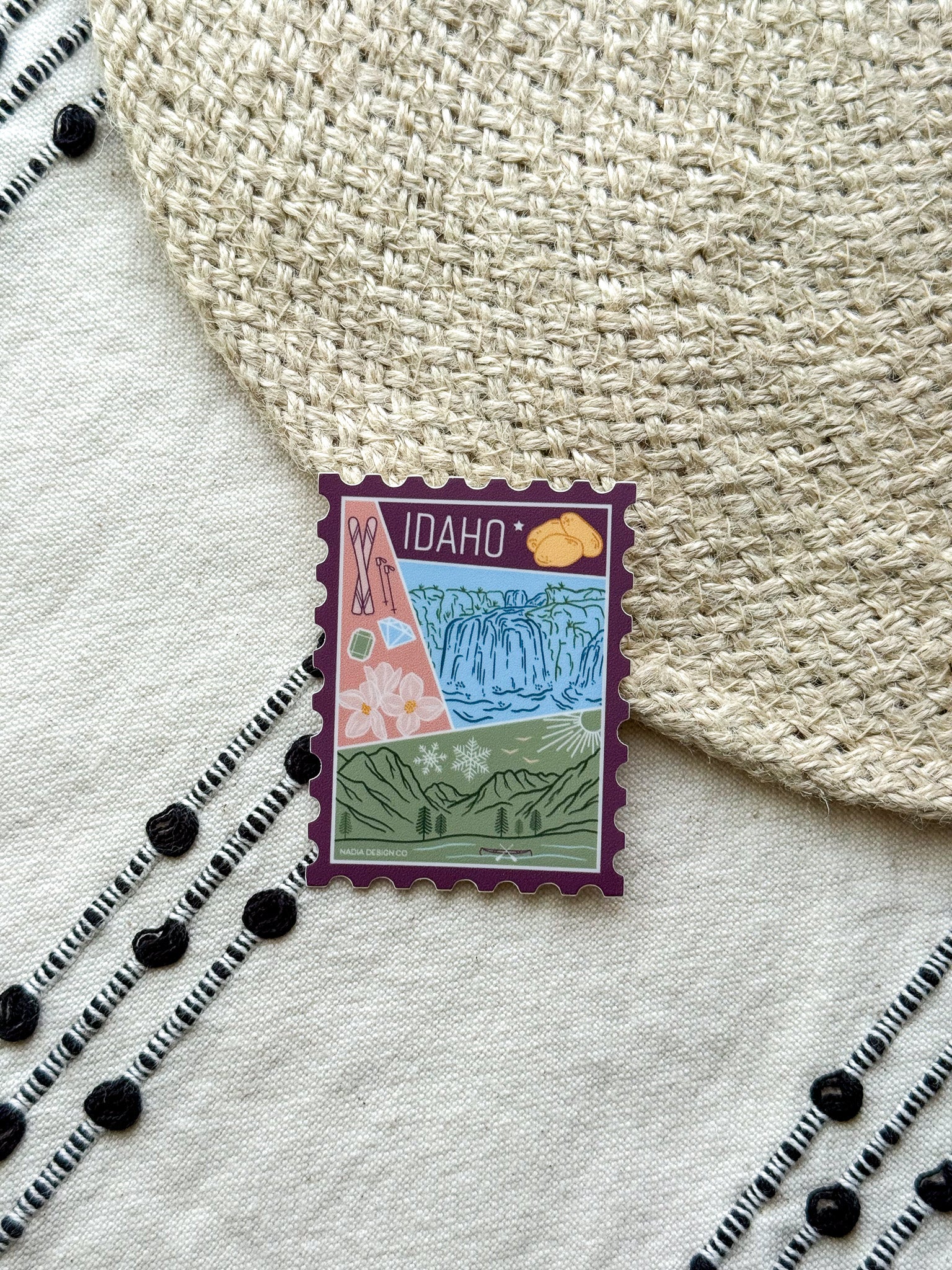 sticker of Idaho with scenic illustrations stamp design held by a hand on a textured fabric background