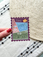 magnet of Idaho with scenic illustrations stamp design held by a hand on a textured fabric background