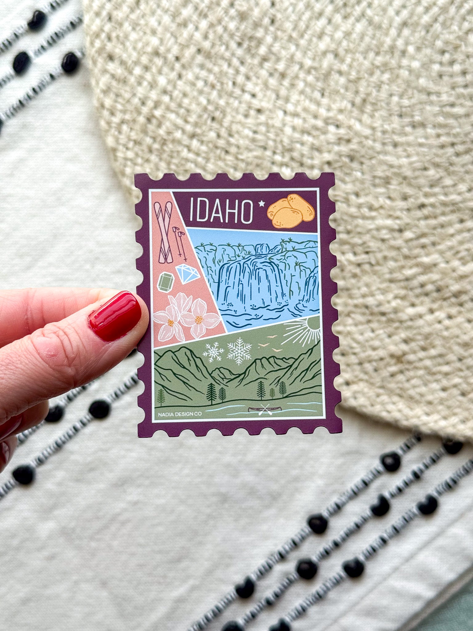 magnet of Idaho with scenic illustrations stamp design held by a hand on a textured fabric background