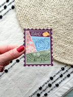 magnet of Idaho with scenic illustrations stamp design held by a hand on a textured fabric background
