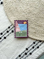 magnet of Idaho with scenic illustrations stamp design held by a hand on a textured fabric background