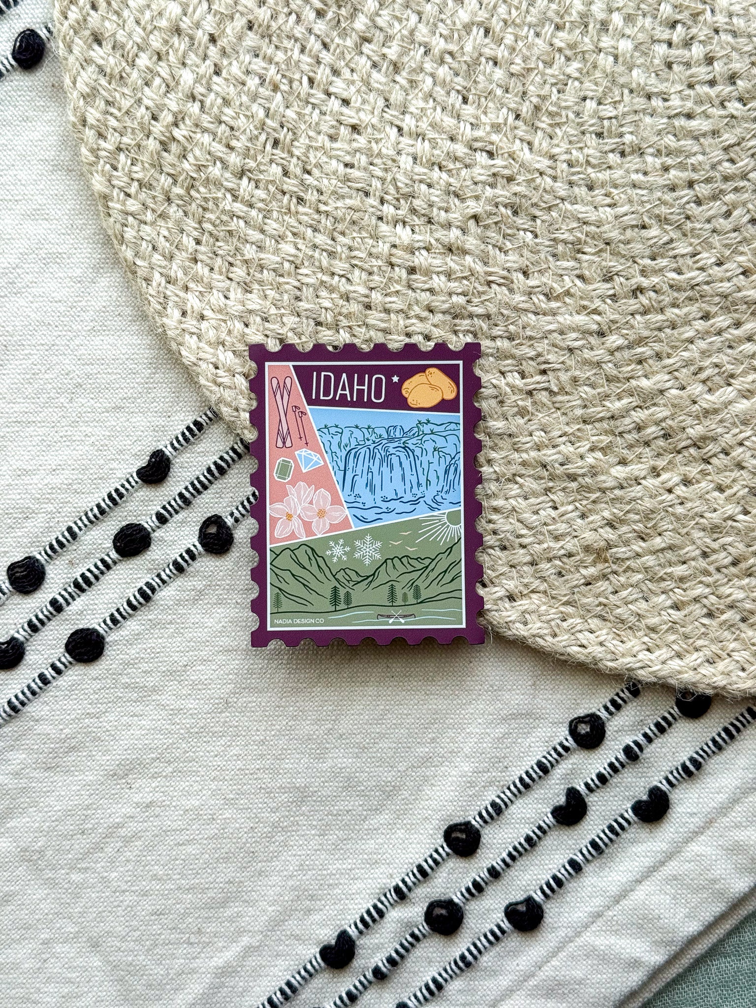 magnet of Idaho with scenic illustrations stamp design held by a hand on a textured fabric background
