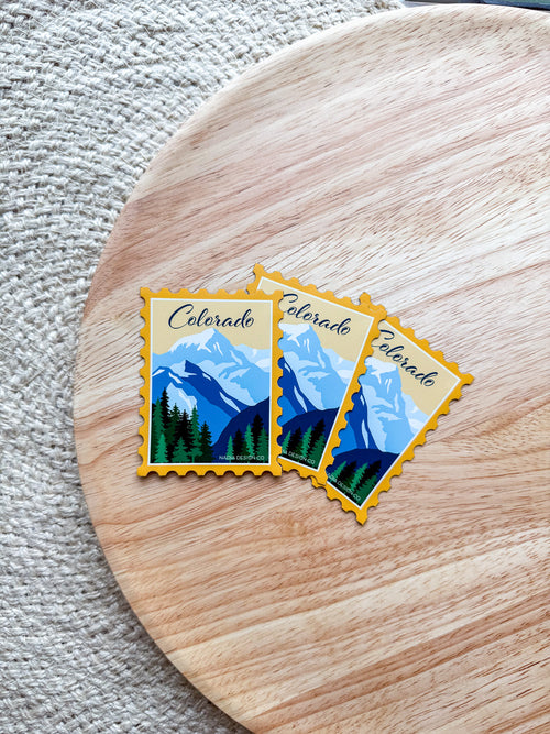 Two Colorado-themed postage stamp magnet designs on a wooden surface.