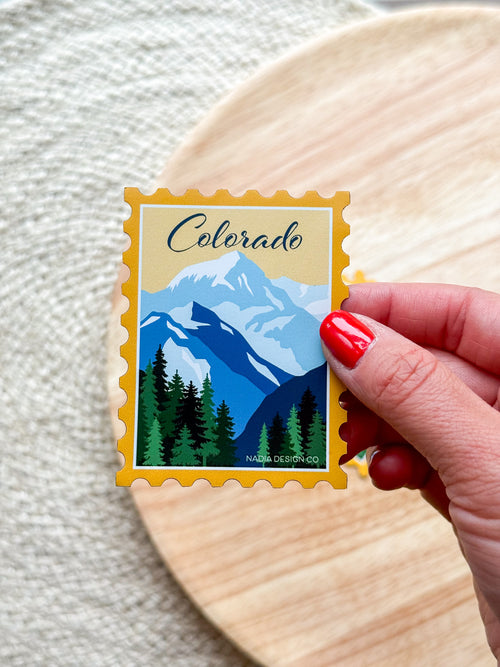 Hand holding a Colorado-themed stamp magnet design on a wooden surface