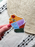 colorful sticker with wisconsin themed illustrations, outline design held by a hand on a textured fabric background