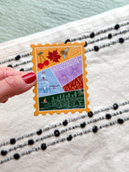 Colorful Wisconsin-themed stamp design held by a hand with red nail polish on a textured surface.
Wisconsin State stamp design sticker with illustration inside with nature and floral themed