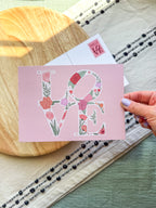 Card with floral 'LOVE' design held by a hand on a wooden surface.