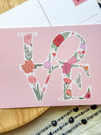 Floral 'LOVE' design on a pink card with a wooden surface and white paper in the background.
