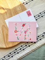 Floral 'LOVE' card on a wooden surface with a patterned background