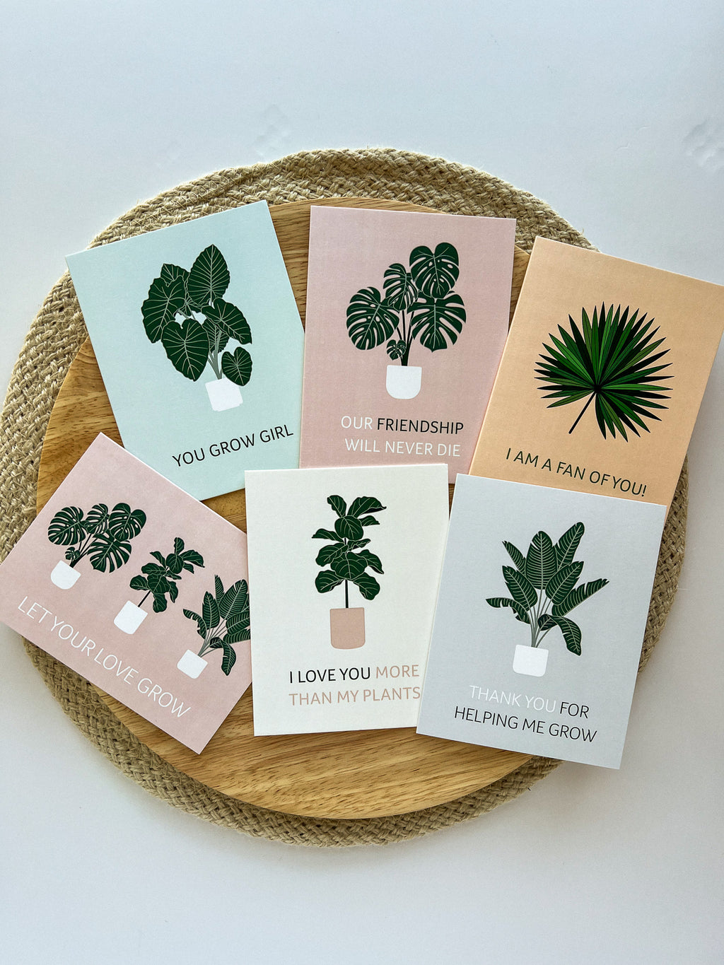 A set of six illustrated greeting cards with various houseplants and punny captions related to plants displayed on a wooden surface.