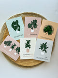 A set of six illustrated greeting cards with various houseplants and punny captions related to plants displayed on a wooden surface.