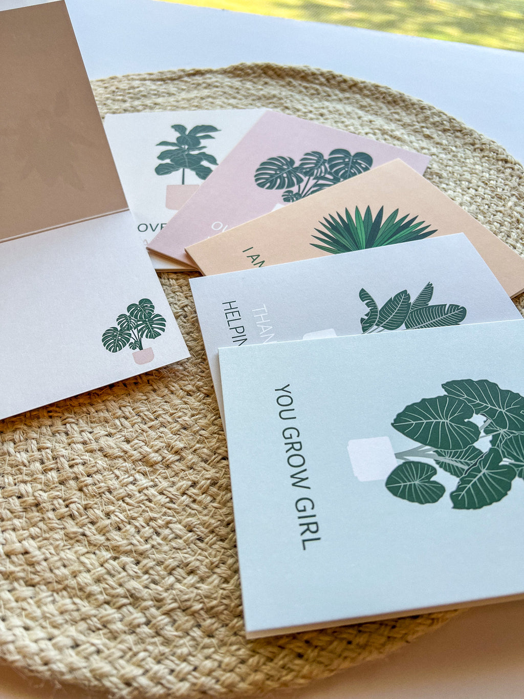 A set of six illustrated greeting cards with various houseplants and punny captions related to plants displayed on a wooden surface.