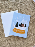 A Merry Christmas card with a snowglobe design featuring a cabin, trees, and snow, placed on a woven surface. Includes a white envelope.
