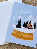 A Merry Christmas card with a snowglobe design featuring a cabin, trees, and snow, placed on a woven surface. Includes a white envelope.
