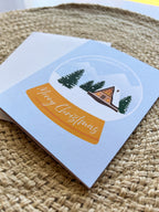 A Merry Christmas card with a snowglobe design featuring a cabin, trees, and snow, placed on a woven surface. Includes a white envelope.