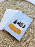 A Merry Christmas card with a snowglobe design featuring a cabin, trees, and snow, placed on a woven surface. Includes a white envelope.