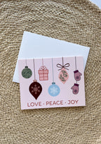 A greeting card with a Christmas design, including the words 'Love Peace Joy' and illustrations of ornaments, a snowflake, and a cabin. The card is placed on a woven matte background.