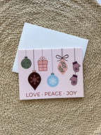 A Christmas themed greeting card with illustrated ornaments and the text 'Merry Christmas' and 'Peace' on a pink background, accompanied by a white envelope, placed on a woven surface.