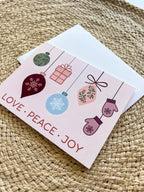 A Christmas themed greeting card with illustrated ornaments and the text 'Merry Christmas' and 'Peace' on a pink background, accompanied by a white envelope, placed on a woven surface.