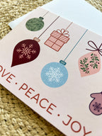 A Christmas themed greeting card with illustrated ornaments and the text 'Merry Christmas' and 'Peace' on a pink background, accompanied by a white envelope, placed on a woven surface.