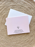 A Christmas themed greeting card with illustrated ornaments and the text 'Merry Christmas' and 'Peace' on a pink background, accompanied by a white envelope, placed on a woven surface.