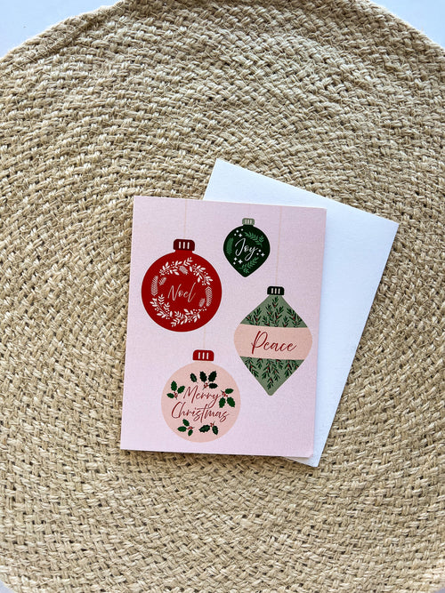 A Christmas themed greeting card with illustrated ornaments and the text 'Merry Christmas' and 'Peace' on a pink background, accompanied by a white envelope, placed on a woven surface.
