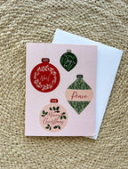 A Christmas themed greeting card with illustrated ornaments and the text 'Merry Christmas' and 'Peace' on a pink background, accompanied by a white envelope, placed on a woven surface.