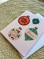 A Christmas themed greeting card with illustrated ornaments and the text 'Merry Christmas' and 'Peace' on a pink background, accompanied by a white envelope, placed on a woven surface.