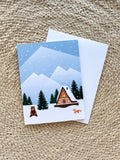 A Christmas greeting card depicting a snowy cabin scene with trees and mountains in the background, a bear, and an orange fox in the foreground. The card is placed next to its white envelope, presented on a woven surface.