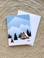 A Christmas greeting card depicting a snowy cabin scene with trees and mountains in the background, a bear, and an orange fox in the foreground. The card is placed next to its white envelope, presented on a woven surface.