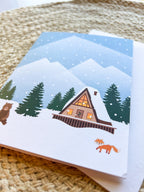A Christmas greeting card depicting a snowy cabin scene with trees and mountains in the background, a bear, and an orange fox in the foreground. The card is placed next to its white envelope, presented on a woven surface.