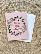 A Christmas greeting card with a floral wreath design and the text 'Oh Come Let Us Adore Him' on a pink background, accompanied by a white envelope, displayed on a woven surface.