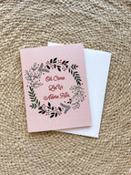 A Christmas greeting card with a floral wreath design and the text 'Oh Come Let Us Adore Him' on a pink background, accompanied by a white envelope, displayed on a woven surface.
