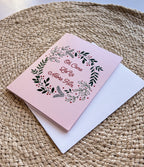 A Christmas greeting card with a floral wreath design and the text 'Oh Come Let Us Adore Him' on a pink background, accompanied by a white envelope, displayed on a woven surface.