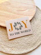A Christmas greeting card with 'JOY TO THE WORLD' written on it, placed on a wooden surface with a white envelope beside it. 