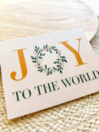 A Christmas greeting card with 'JOY TO THE WORLD' written on it, placed on a wooden surface with a white envelope beside it. 
