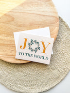 A Christmas greeting card with 'JOY TO THE WORLD' written on it, placed on a wooden surface with a white envelope beside it. 