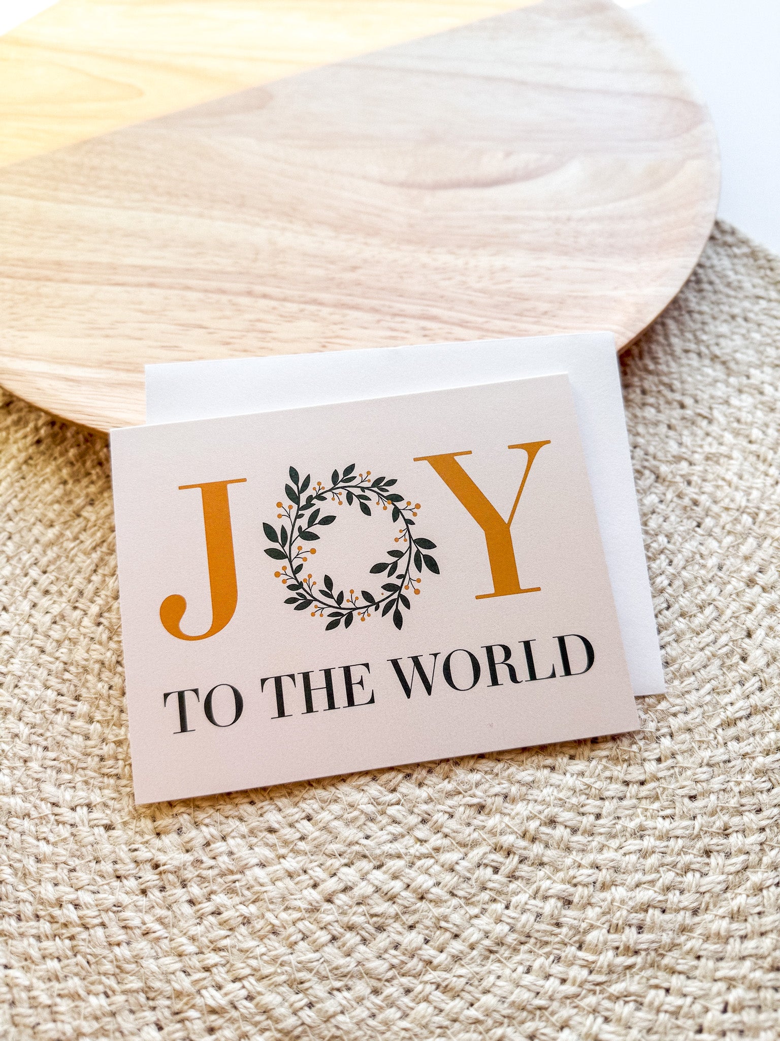A Christmas greeting card with 'JOY TO THE WORLD' written on it, placed on a wooden surface with a white envelope beside it. 