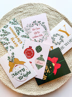 A set of six Christmas-themed greeting cards with envelopes, displayed on a round, beige textured mat. The cards feature various designs including 'Merry Christmas' with ornaments, 'Joy to the World' with a Christmas tree, and other festive images.