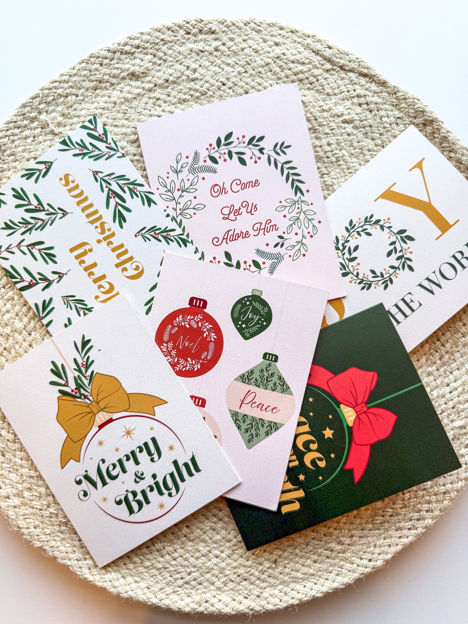 A set of six Christmas-themed greeting cards with envelopes, displayed on a round, beige textured mat. The cards feature various designs including 'Merry Christmas' with ornaments, 'Joy to the World' with a Christmas tree, and other festive images.