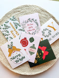 A set of six Christmas-themed greeting cards with envelopes, displayed on a round, beige textured mat. The cards feature various designs including 'Merry Christmas' with ornaments, 'Joy to the World' with a Christmas tree, and other festive images.