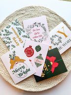 A set of six Christmas-themed greeting cards with envelopes, displayed on a round, beige textured mat. The cards feature various designs including 'Merry Christmas' with ornaments, 'Joy to the World' with a Christmas tree, and other festive images.
